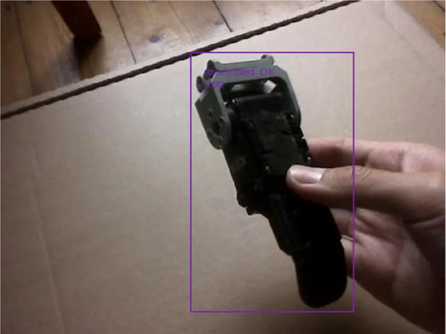 Hand holding a black plastic part in front of the camera, detected inside a bounding box